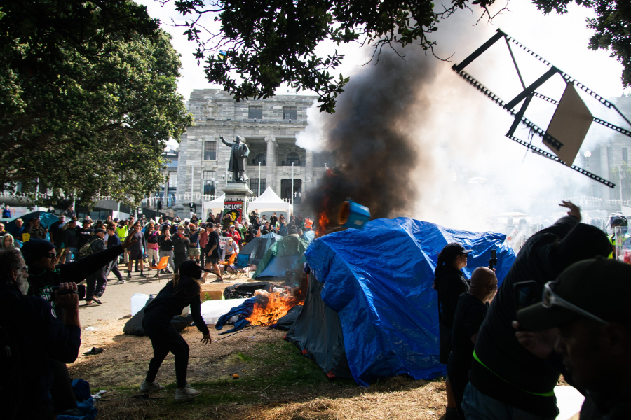 A tent burning on New Zealand's Parliament grounds with a plume of black smoke. Protesters look on.